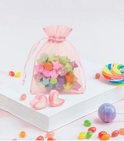 Sachet en organza rose translucide avec cordon de serrage, posé sur un livre blanc et rempli de petites étoiles en papier colorées.